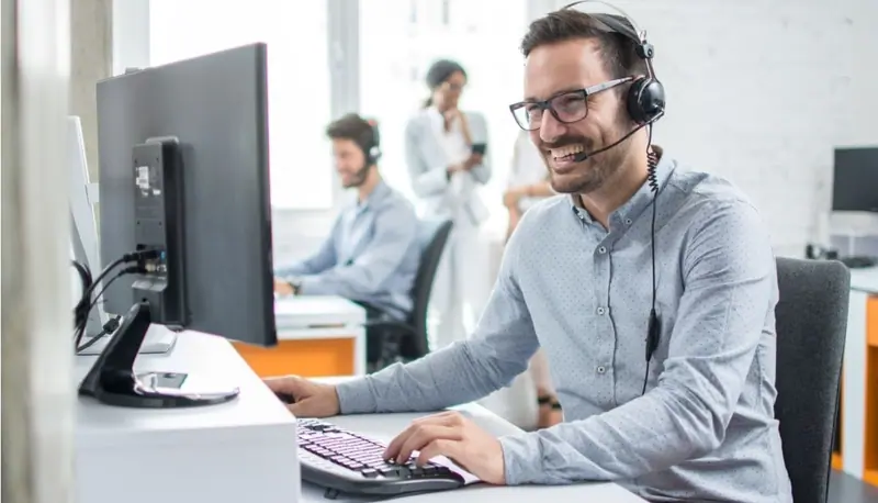 Smiling customer service agent with a headset typing at a computer in a bright office